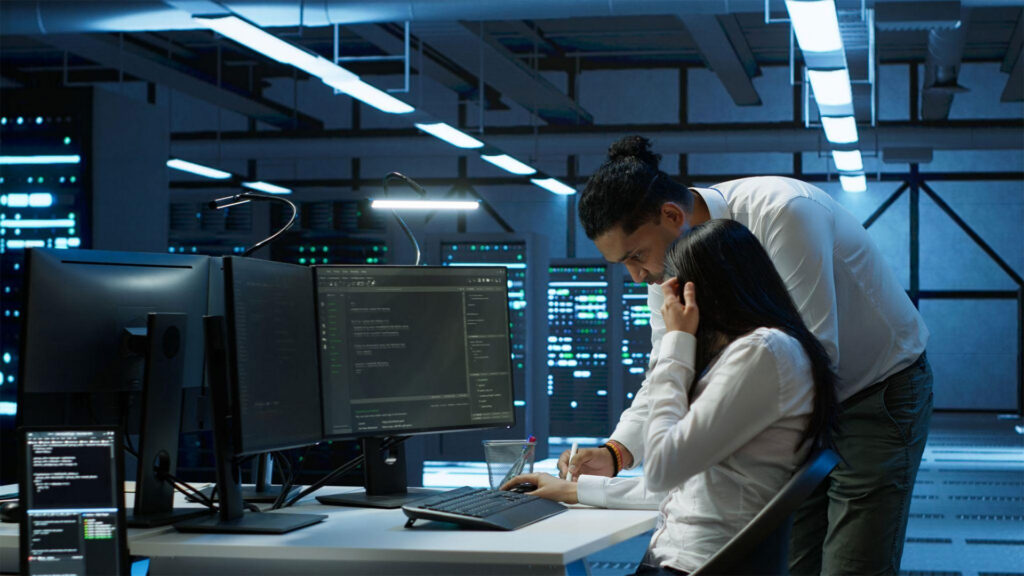 Two IT professionals work at a dual-monitor workstation in a server room, reviewing code on screens under blue lighting.