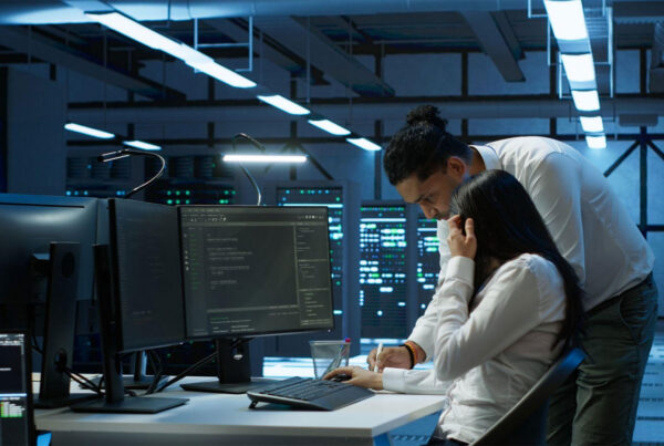 Two IT professionals work at a dual-monitor workstation in a server room, reviewing code on screens under blue lighting.